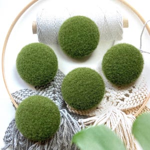 Op de afbeelding: Vijf ronde, pluizige, olijfgroene vilten knopen gerangschikt in een ronde houten borduurring. Onder de knopen liggen verschillende lengtes wit en grijs katoenen koord, waarvan sommige voorzien zijn van macramé knopen en franjes.