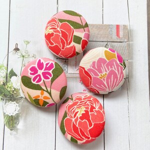 May include: Four circular fabric-covered buttons featuring vibrant floral patterns in shades of red, pink, green, and orange. Each button displays a distinct botanical print with stylized flowers and leaves on a textured, woven textile surface.
