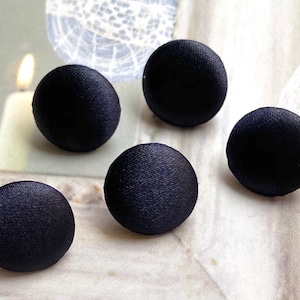 Könnte beinhalten: Fünf schwarze Satin-Knöpfe, jeder mit einer glatten, leicht strukturierten Oberfläche. Die Knöpfe sind in einem Halbkreis auf einer weißen Oberfläche angeordnet.