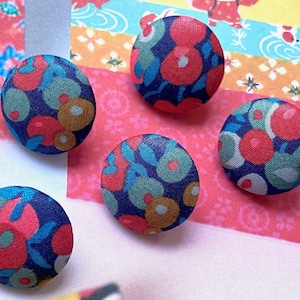 Puede incluir: Cinco botones redondos cubiertos de tela con un estampado floral. Los botones presentan un fondo azul marino con diseños florales rojos, turquesa y amarillo-marrón. Están colocados sobre un fondo colorido con patrones.