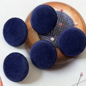 May include: Four circular buttons covered in a deep navy blue velvet fabric. The buttons have a smooth, rounded surface and a soft, fuzzy texture. They are arranged around a wooden pincushion that features a grey herringbone fabric center.