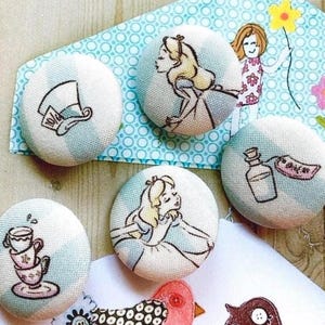 Peut inclure: Un ensemble de cinq boutons ronds recouverts de tissu avec des illustrations d'Alice au pays des merveilles, dont un haut-de-forme, une fille aux cheveux blonds, une bouteille en verre avec une étiquette et une pile de tasses à thé.