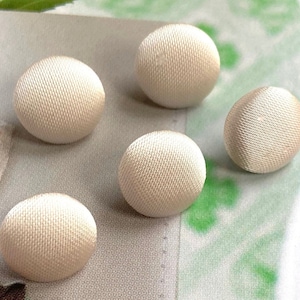 Op de afbeelding: Vijf ronde knopen bekleed met een gladde, gebroken witte satijnen stof. Elke knoop heeft een bolle, convexe vorm met een zichtbare, fijn geweven textuur over het oppervlak. De knopen zijn verspreid over een plat oppervlak gerangschikt.