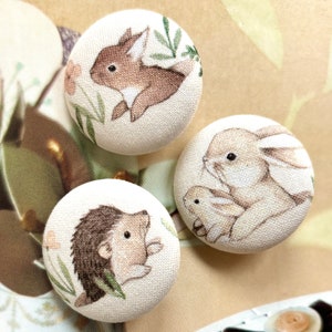 Peut inclure: Trois boutons ronds recouverts de tissu avec des illustrations imprimées d'animaux des bois. Un bouton montre un lapin brun, un autre un hérisson, et le troisième représente un grand lapin avec un petit lapereau, sur un fond crème.