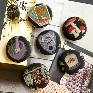 May include: Six circular fabric-covered buttons featuring black backgrounds and various printed designs including a tarot card labelled The Sun, red mushrooms, a mortar and pestle with herbs, a planchette, a moth in a glass cloche, and pine branches.