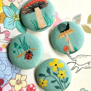 Peut inclure: Quatre boutons ronds recouverts de tissu bleu sarcelle clair. Chaque bouton présente un motif brodé différent : un grand champignon, un escargot sur un champignon, une coccinelle avec de l'herbe verte et des fleurs jaunes à feuilles vertes.