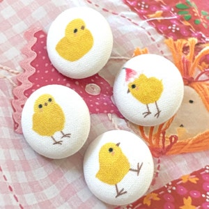 May include: Four white fabric-covered buttons with embroidered yellow chicks. One chick wears a Santa hat. The buttons are round and have a textured surface. The background is a pink and white patchwork pattern.