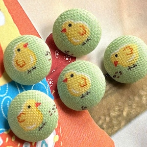 May include: Five round fabric buttons with a light green background and a yellow chick design. The chicks have red beaks and black eyes. The buttons are likely for sewing or crafts.
