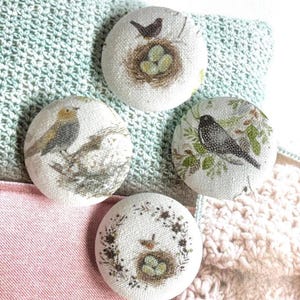 Peut inclure: Quatre boutons ronds recouverts de tissu avec des illustrations imprimées. L'un montre un oiseau au-dessus d'un nid avec des œufs, un autre un oiseau sur une branche, un troisième un oiseau parmi des feuilles vertes et le quatrième un oiseau.