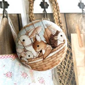 May include: A circular fabric button with a detailed illustration of squirrels and rabbits nestled in a woven basket. The button has a light blue background with a floral pattern and is attached to a jute rope.