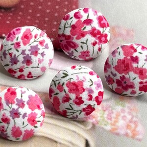 May include: Five round fabric-covered buttons with a floral pattern. The buttons feature a white background with a repeating pattern of pink and purple flowers. The buttons are arranged on a light background.