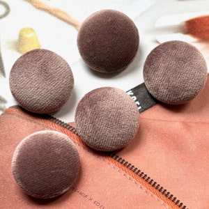 Peut inclure: Cinq boutons circulaires recouverts d'un tissu en velours mauve doux. Chaque bouton présente une forme bombée lisse et arrondie avec une texture pelucheuse et uniforme sur toute la surface, disposés en groupe pour montrer la finition.
