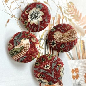 Pode incluir: Quatro botões redondos de tecido com um padrão floral e de pássaros em tons de bordô, creme e verde. Os botões estão dispostos sobre uma superfície branca com um fundo neutro.