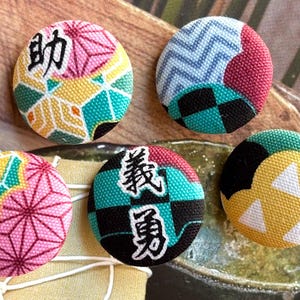 Pode incluir: Cinco botões circulares cobertos de tecido com vários padrões e cores. Dois botões exibem caracteres kanji japoneses pretos. Os padrões incluem designs geométricos em rosa e amarelo, uma estampa de chevron azul e um motivo de triângulos amarelos.