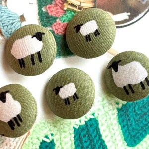 Pode incluir: Cinco botões redondos cobertos de tecido verde-oliva com um desenho de ovelha branca. Cada botão apresenta uma ovelha branca com cabeça e patas pretas. Os botões são redondos e têm uma aparência texturizada, adequados para costura ou artesanato.