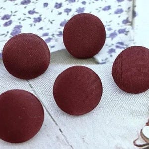 May include: Five circular buttons covered in a deep burgundy fabric with a smooth, matte finish. Each button features a simple, rounded dome shape with a consistent texture across the surface, arranged in a cluster to display the fabric color.