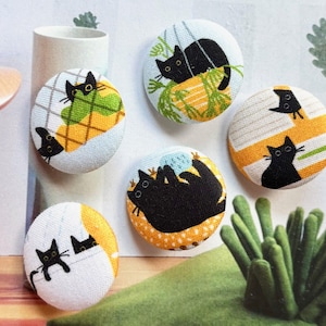 Op de afbeelding: Een verzameling van zes ronde stoffen knopen, elk met een illustratie van een zwarte kat. De katten worden in verschillende poses afgebeeld tegen achtergronden met patronen in geel, groen en wit. Een charmant knutselartikel.