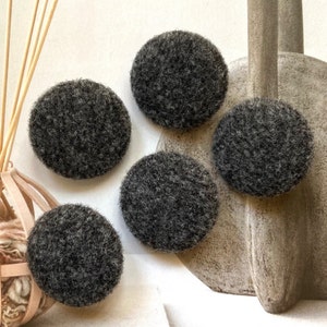 Puede incluir: Cuatro botones circulares con una superficie de lana fieltrada texturizada en gris oscuro. Cada botón presenta una apariencia densa y fibrosa con variaciones sutiles en las fibras de color carbón, creando una forma redondeada y uniforme.