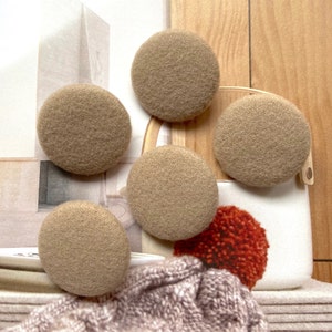 Op de afbeelding: Vijf beige, ronde vilten pads worden getoond. De pads hebben een zacht, getextureerd uiterlijk. Een witte container met een houten handvat en een rode pompon staan op de achtergrond. De pads worden waarschijnlijk gebruikt voor meubelbescherming.