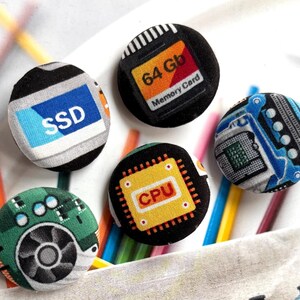 Op de afbeelding: Vier ronde, met stof beklede knopen met technologie-prints. Eén knop toont een blauw SSD-label, een andere een 64 Gb geheugenkaart, een derde een gele CPU-chip en de vierde een groene printplaat met een afbeelding van een koelventilator.