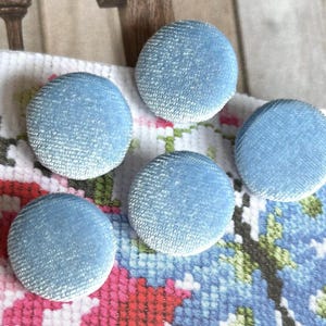 May include: Five round, pale blue fabric-covered buttons. The buttons are placed on a colourful cross-stitch pattern with pink, red, green, and blue floral designs. The buttons have a textured appearance.