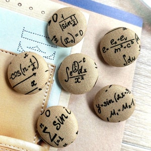 Pode incluir: Seis botões redondos cobertos de tecido bege com equações e símbolos matemáticos pretos. Os botões estão dispostos sobre uma superfície castanha clara, com um caderno no fundo.