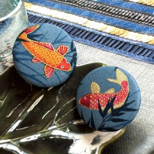 Può includere: Due bottoni rotondi in tessuto con sfondo blu e design di pesce koi. I pesci sono arancioni e rossi con pinne e squame dettagliate. I bottoni sono su un piatto a forma di foglia.