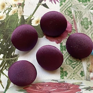 May include: Four circular buttons covered in a deep purple fabric with a visible satin-like sheen and a fine, diagonal ribbed texture. Each button has a smooth, domed surface and a consistent, solid dark plum color throughout the set.