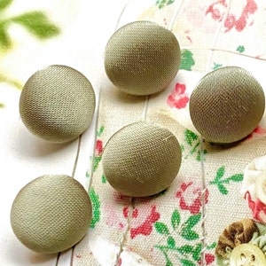 May include: Five olive-green fabric-covered buttons. The buttons are round with a textured surface. The buttons are arranged on a floral patterned fabric background.