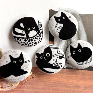 May include: Five circular fabric buttons with black cat illustrations. The cats are shown in different poses, with white details on a light grey backdrop. The buttons showcase a blend of playful and minimalist designs.