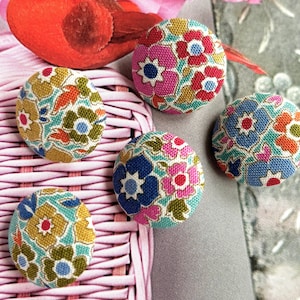 May include: Four circular fabric-covered buttons featuring a floral pattern. The buttons display various color combinations including pink, blue, yellow, green, and orange flowers on a light background. The buttons are arranged on a pink woven surface.