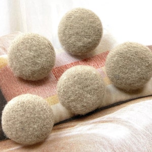 以下が含まれることがあります： ベージュのフェルトボールが5つ、模様入りの布の上に配置されています。ボールは丸く、ふわふわした質感です。布は茶色、オレンジ、白などの色合いです。各ボールの直径は約4cmです。
