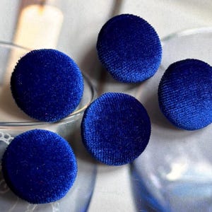 May include: Five round, cobalt blue velvet buttons. The buttons are arranged on a light background, with a soft, textured appearance. These buttons are suitable for sewing projects, crafts, or adding a touch of elegance to garments.