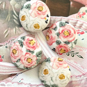 Puede incluir: Cuatro botones circulares cubiertos de tela con un estampado floral de flores rosas y blancas, centros amarillos y hojas verdes sobre un fondo crema. Los botones están dispuestos en grupo, mostrando el diseño textil consistente.