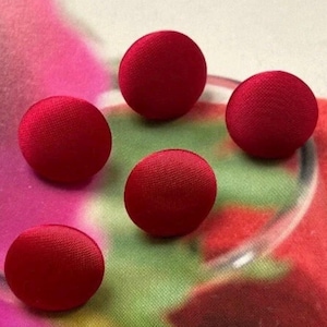 Può includere: Cinque bottoni rotondi rossi rivestiti in tessuto sono sparsi su uno sfondo colorato. I bottoni hanno una forma leggermente bombata e una superficie liscia e strutturata. Lo sfondo presenta una miscela di tonalità rosa, verdi e bianche.