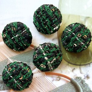 Op de afbeelding: Vijf ronde tweed knopen met een zwart-groen weefpatroon en gouden en witte accenten. De knopen liggen op een wit oppervlak, met een groene glazen vaas op de achtergrond. Elke knoop heeft een diameter van ongeveer 2,5 cm.