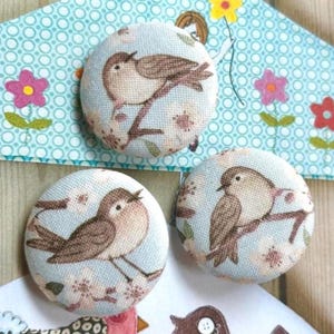 Peut inclure: Trois boutons ronds recouverts de tissu avec un fond bleu clair et un motif répétitif d'oiseaux bruns perchés sur des branches, agrémentés de petites fleurs roses et blanches. Les boutons ont une surface lisse et arrondie.