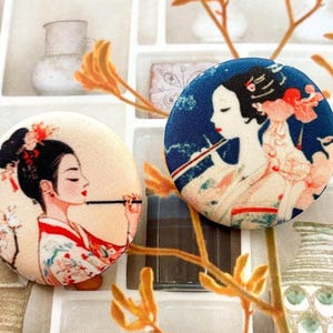 Può includere: Due bottoni decorativi rotondi con illustrazioni di geishe. Un bottone ha uno sfondo color crema con una geisha in kimono rosso e bianco. L'altro bottone ha uno sfondo blu con una geisha che suona il flauto.