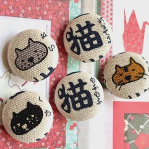 May include: Five circular fabric-covered buttons featuring beige linen-like material. Three buttons display stylised cat faces in black, grey, and orange. Two buttons feature the black Japanese kanji character for cat, 猫, with small hiragana text.