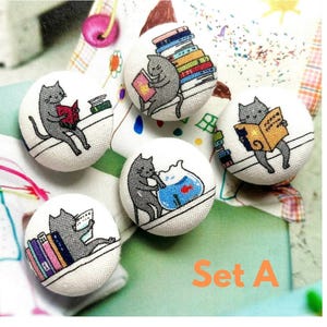 May include: A set of five white fabric buttons with embroidered cat designs. Each button shows a grey cat reading a book or interacting with a fishbowl. The text "Set A" is printed in orange. These buttons are suitable for crafting and sewing projects.