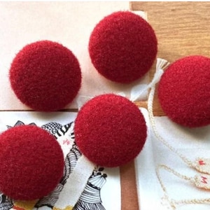 Può includere: Quattro sfere rotonde in lana infeltrita rosso brillante con una superficie ruvida e testurizzata. Ogni sfera è attaccata a una sottile striscia di tessuto intrecciato bianco sporco con una piccola lettera M rossa stampata sull'estremità.