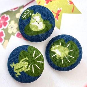 Könnte beinhalten: Drei runde Stoffknöpfe mit blauem Denim-Hintergrund. Jeder Knopf zeigt einen grünen Frosch auf einem Seerosenblatt. Ein Frosch hält ein Blatt, die anderen sind in verschiedenen Posen. Ein skurriles Accessoire.