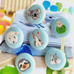 May include: A selection of handmade fabric buttons, each with a different animal face. The buttons feature a koala, sheep, rabbit, sloth, hamster, and parakeet, on light blue fabric. Ideal for sewing and craft projects.