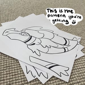 Puede incluir: Un patrón imprimible para una manualidad de dragón. El patrón está impreso en papel blanco y muestra un dragón con alas, cola y cabeza. El texto "This is the pattern you're getting" está escrito en el papel.