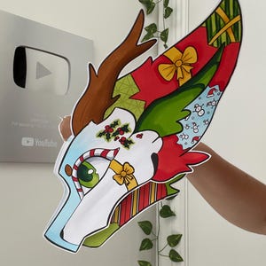May include: A colorful, cartoonish animal head with Christmas-themed decorations. The design features red, green, and blue sections with gift boxes, candy canes, and holly. The head has brown antlers and a green eye.