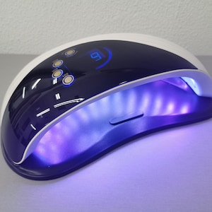 Könnte beinhalten: Eine weiße und marineblaue UV-Nagellampe mit digitaler Zeitanzeige. Die Lampe hat drei Timer-Tasten und strahlt violettes Licht aus. Das Design ist schlank und modern, geeignet für den professionellen oder privaten Gebrauch.