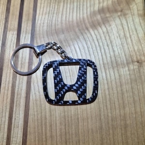 May include: A black and white carbon fiber-style keychain featuring the Honda logo. The keychain has a silver-colored ring and chain. The background is a wooden surface with vertical grain.