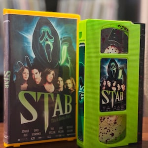May include: Two VHS tapes with the movie title "STAB". One tape has a black cover with a ghost face and the movie title. The other tape is bright green with a similar design and blood splatter.
