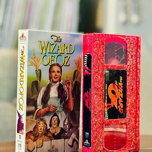 May include: Two VHS tapes of "The Wizard of Oz". One tape has a colourful cover with Dorothy and the characters. The other tape is red with glitter and the movie title. The tapes are side by side.