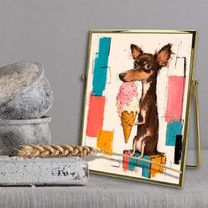 K&ouml;nnte beinhalten: Ein gerahmter Kunstdruck, der einen braunen Hund mit gro&szlig;en Ohren zeigt, der eine rosa Eist&uuml;te h&auml;lt und auf einem bunten Block sitzt. Der Hintergrund besteht aus abstrakten rechteckigen Formen in Blau-, Rosa-, Orange- und Schwarzt&ouml;nen auf Creme.